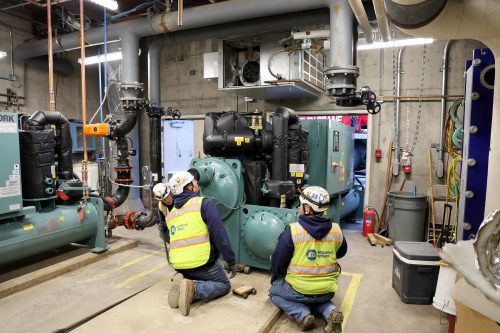 Technicians in hard hats and safety gear retrofitting a large chiller into an HVAC system. The chiller, a crucial component of the HVAC setup, is carefully positioned by the workers as part of maintenance service for this government project in Tucson, Arizona.